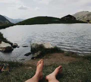 barefooted person lying on grass viewing lake and mountain